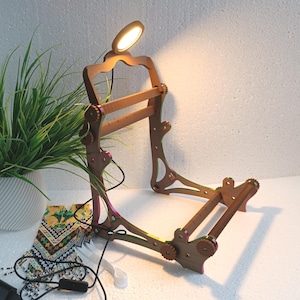 May include: A brown wooden desk lamp with an adjustable frame and a warm light. The lamp has a small, round light at the top. A green plant in a white pot and a decorative pouch are also in the image.