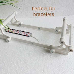 May include: A white adjustable bracelet display stand with a bracelet. The stand has a rectangular frame with adjustable arms and a series of rollers. The bracelet is black with a floral pattern and a silver chain.