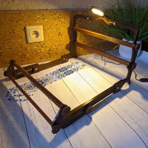 Peut inclure: Un métier à broder en bois marron foncé avec une lampe LED réglable intégrée. Le cadre maintient un morceau de tissu avec un motif bleu et jaune. La lampe est allumée, éclairant le cadre et le tissu.