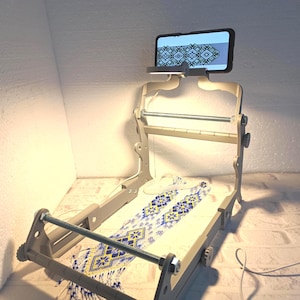 Könnte beinhalten: Ein hölzerner Stickrahmen mit einem Smartphone, das ein Muster anzeigt. Ein besticktes Stück mit blauen und gelben floralen Designs befindet sich im Rahmen. Das Setup wird von einem Licht beleuchtet, was auf ein Bastel- oder DIY-Projekt hindeutet, etwa 60 cm lang.
