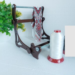 May include: A wooden bead loom with a red and white beaded necklace on it. A spool of white thread with a red base is in the foreground.