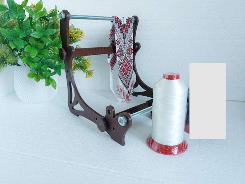 May include: A wooden bead loom with a red and white beaded necklace on it. A spool of white thread with a red base is in the foreground.