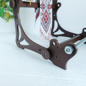 May include: A brown wooden loom with a metal rod and a white and red patterned fabric hanging from it.