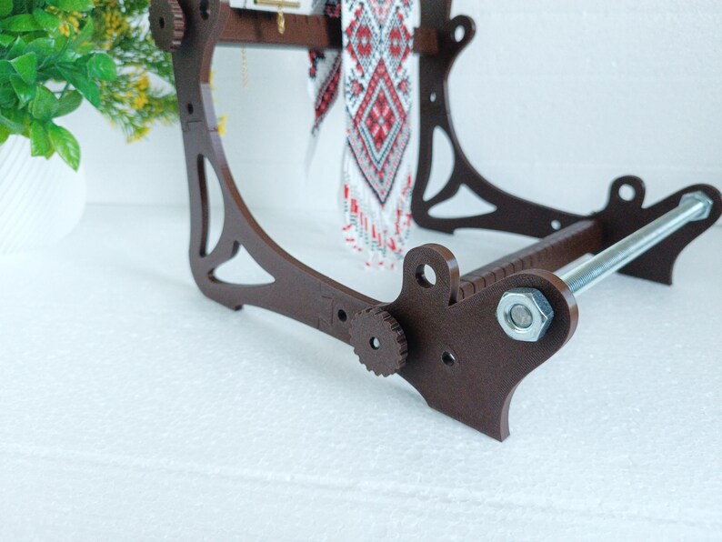 May include: A brown wooden loom with a metal rod and a white and red patterned fabric hanging from it.