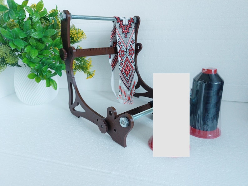 May include: A wooden bead loom with a red and white beaded necklace on it. A spool of black thread is on the right side of the image.
