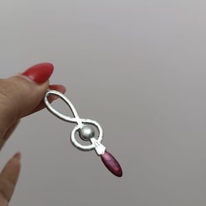 Peut inclure: Pendentif argenté en forme de clé de sol. Il comporte une petite perle sphérique et un détail en forme de goutte de couleur bordeaux. Le pendentif est tenu par une main avec du vernis à ongles rouge.