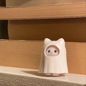 Puede incluir: Una pequeña figura blanca de un fantasma con orejas de gato. El fantasma tiene una abertura redonda que revela un pequeño animal peludo con ojos negros. La figura está colocada sobre un fondo de libros apilados de color marrón claro.