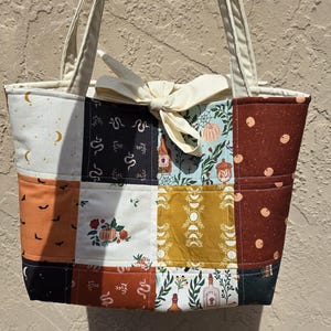 May include: A patchwork tote bag featuring a variety of fabric squares with patterns including pumpkins, snakes, moons, flowers, and bottles. The bag has two long cream-colored shoulder straps and a cream-colored fabric tie closure at the top center.