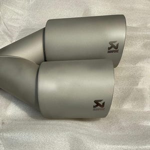 Puede incluir: Un par de salidas de escape plateadas con un logotipo Akrapovic negro. Las salidas están conectadas por un tubo plateado.