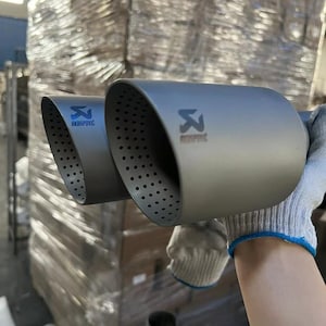 Puede incluir: Un par de puntas de escape grises con el logotipo de Akrapovic. Las puntas tienen un diseño perforado y están sujetas por una mano enguantada. El fondo muestra paquetes envueltos.