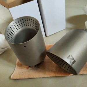 May include: Two silver-colored exhaust tips with perforated ends. One tip is upright, revealing its interior, while the other lies on its side, displaying the Akrapovic logo. They rest on a small, tan towel.