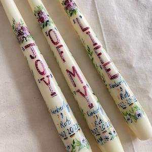Könnte beinhalten: Drei elfenbeinfarbene Kerzen mit handgemalten Blumendesigns und den Worten "JOY OF LIFE" in burgunderfarbener Schrift. Jede Kerze ist mit einem Namen und Datum personalisiert und hat einen floralen Rand in Lila, Grün und Weiß.