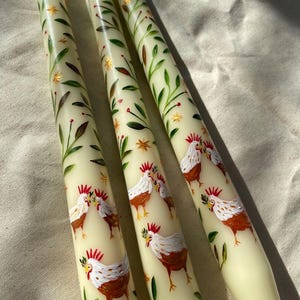 May include: Three ivory-colored taper candles with hand-painted designs. Each candle features a repeating pattern of chickens, green leaves, and gold stars. The candles are arranged diagonally, showcasing the detailed artwork.