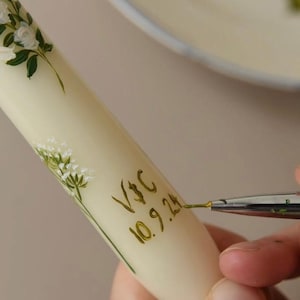 Op de afbeelding: Een witte kaars met groene bloemenmotieven en gouden letters. De letters zeggen "V+C 10.9.24".