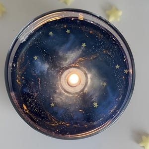 Könnte beinhalten: Eine schwarze und blaue Galaxie-Kerze mit goldenem Glitzer und einer kleinen weißen Flamme in der Mitte. Die Kerze ist von kleinen goldenen Sternen umgeben.