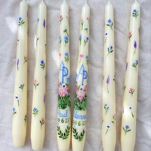 May include: Six white taper candles with floral designs and personalized names. The candles are decorated with blue, pink, and purple flowers. The names on the candles are Paul, Ianna, and AP. The candles are on a white background.