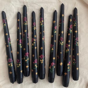 Könnte beinhalten: Zehn schwarze Stumpenkerzen mit einem floralen Design. Die Kerzen sind mit roten und gelben Blumen und goldenen Sternen verziert.