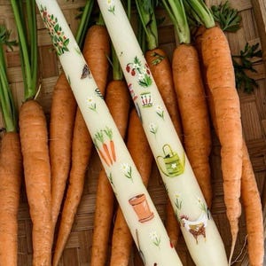 Könnte beinhalten: Zwei weiße Kerzen mit handgemalten Motiven von Gartengeräten, Blumen und einem Huhn. Die Kerzen werden in einem Weidenkorb mit Karotten präsentiert.