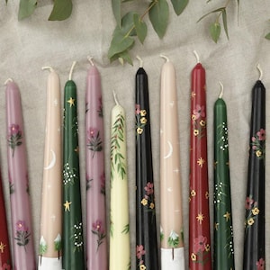 Puede incluir: Colección de velas cónicas pintadas a mano en varios colores, incluyendo rojo, rosa, verde, negro y crema. Cada vela presenta diseños florales, celestiales y botánicos. Las velas están dispuestas sobre una superficie de lino neutra.