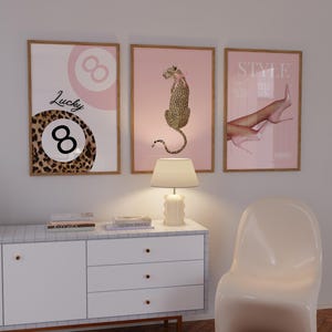 May include: Three framed prints with a pink background. The left print features a black and brown leopard print with a white number eight and the text "Lucky 8". The middle print features a pink leopard sitting on a pink background. The right print features a pair of legs with pink high heels and the text "Style".