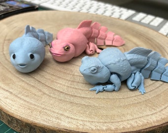 May include: Three small, 3D printed, articulated figures of fantastical creatures. One is pink, one is light blue, and the other is a darker blue. They are posed on a wooden surface.