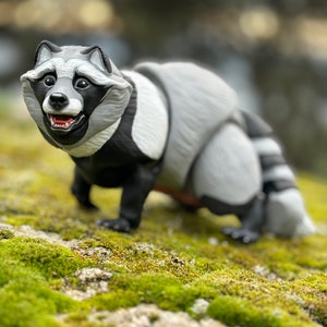 May include: A grey and black raccoon figurine with a playful expression, standing on a bed of green moss.