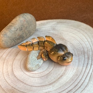 May include: A hand-painted ceramic figurine of a brown and gold frog with large eyes. The frog is sitting on a wooden surface with a small rock beside it.
