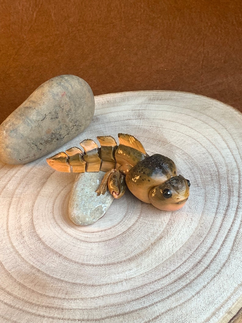 May include: A hand-painted ceramic figurine of a brown and gold frog with large eyes. The frog is sitting on a wooden surface with a small rock beside it.