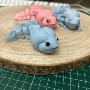 May include: Three small, hand-painted figurines of aquatic creatures on a wooden base. The figurines are painted in shades of blue and pink. The base is made of natural wood with a rough texture.