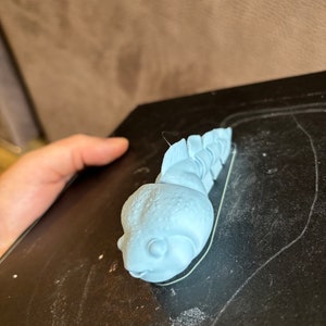 May include: A light blue 3D printed figurine of a small, cartoon-like creature with a large head and a segmented body. The creature has a large, round eye and a small mouth.