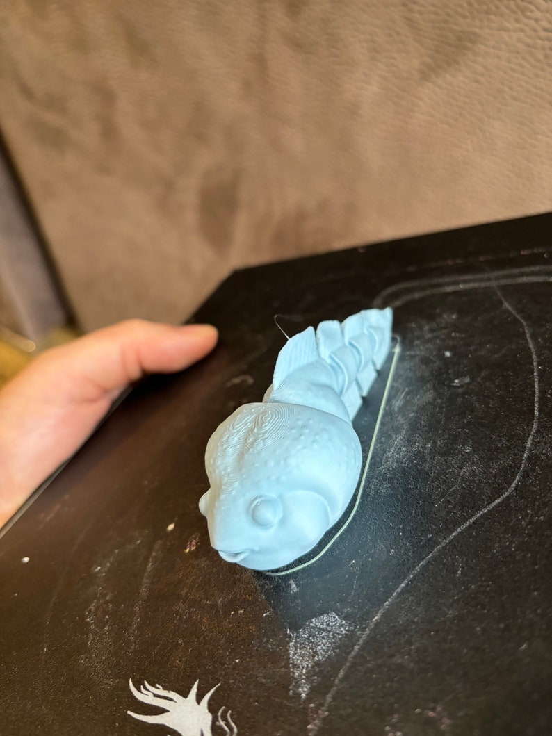 May include: A light blue 3D printed figurine of a small, cartoon-like creature with a large head and a segmented body. The creature has a large, round eye and a small mouth.