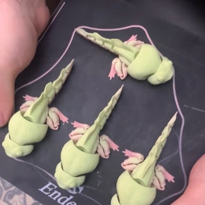 May include: Four 3D printed green axolotl figurines with pink gills on a black surface with the word "Ender" printed on it.