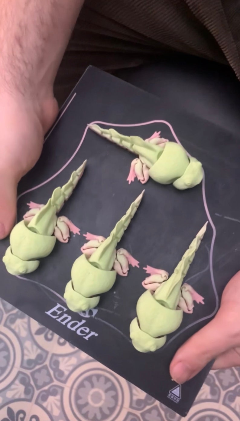 May include: Four 3D printed green axolotl figurines with pink gills on a black surface with the word "Ender" printed on it.