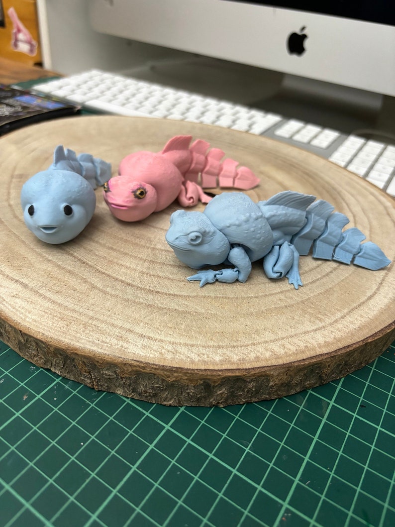 May include: Three small, 3D printed, articulated figures of fantastical creatures. One is pink, one is light blue, and the other is a darker blue. They are posed on a wooden surface.