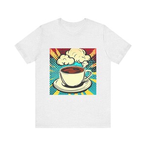 Può includere: Una maglietta bianca con una grafica retrò di una tazza di caffè con vapore che sale da essa. La grafica è su uno sfondo colorato con uno schema di colori blu, giallo e rosso.
