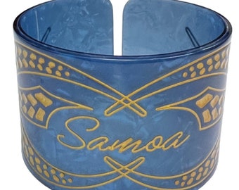 Puede incluir: Una pulsera de plástico azul con un diseño dorado y la palabra "Samoa" escrita en cursiva.