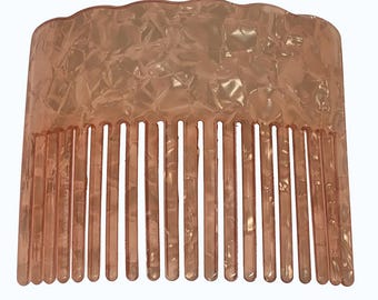 May include: A peach-colored hair comb with a wavy top edge and a series of vertical teeth. The comb has a marbled, textured appearance, and is made of a translucent material.