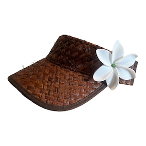 Puede incluir: Una visera tejida marrón con un acento de flor blanca. La visera está hecha de un material tejido y tiene una visera marrón.