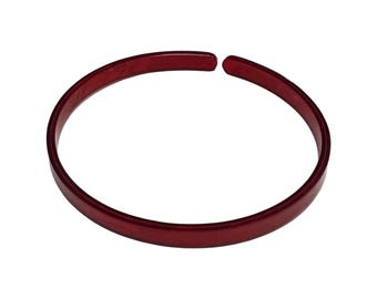 Puede incluir: Una pulsera de puño abierta de color rojo. La pulsera es de un color rojo intenso y sólido, con una superficie lisa y pulida. El diseño es un círculo simple y elegante que no se cierra por completo, dejando un pequeño espacio.