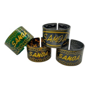 Puede incluir: Cuatro anillos de servilleta decorativos con la palabra "SAMOA" escrita en letras doradas. Los anillos están hechos de diferentes materiales, incluyendo verde, negro y marrón.