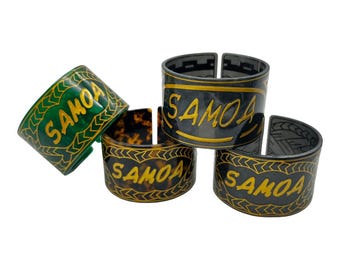 May include: Four decorative napkin rings with the word "SAMOA" written in gold lettering. The rings are made of different materials, including green, black, and brown.