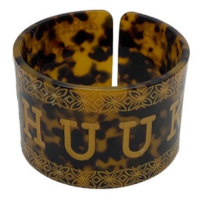 Può includere: Un tovagliolo con anello a motivo marrone e nero con una scritta dorata che recita "HUUK".