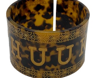 May include: A brown and black patterned napkin ring with a gold inscription that reads "HUUK".