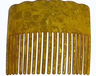 Puede incluir: Un peine para el cabello de color amarillo ámbar translúcido con un patrón moteado similar al carey. El peine presenta un borde superior curvo y festoneado, con una serie de catorce dientes largos, delgados y espaciados uniformemente.
