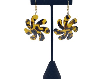 May include: A pair of gold tone dangle earrings with a large, brown and yellow swirl design. The earrings are on a black display stand.