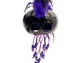 May include: A handcrafted, black coconut mask with round eye holes and purple feather accents. The mask features dangling purple beaded strands and is decorated with black and purple feathers. This unique mask is perfect for costume parties or decorative display.
