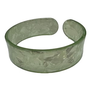 Può includere: Un bracciale a fascia largo e aperto di colore verde salvia pallido traslucido. Il materiale presenta un motivo testurizzato e marmorizzato su tutta la superficie, creando un aspetto sottile e stratificato. Il bracciale ha un bordo arrotondato.