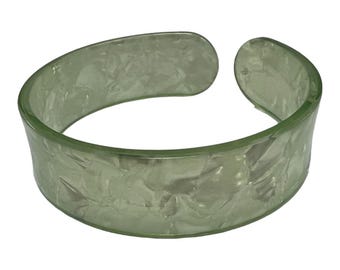 Puede incluir: Una pulsera tipo brazalete ancha y abierta de color verde salvia pálido translúcido. El material presenta un patrón texturizado y jaspeado en toda la superficie, creando una apariencia sutil y estratificada. La pulsera tiene un borde redondeado.