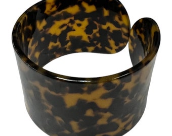 May include: A wide, brown and black tortoiseshell bangle bracelet. The bracelet is made of plastic and has a smooth, glossy finish.