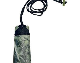 Puede incluir: Un colgante de piedra verde y gris con un cordón trenzado negro. El colgante es rectangular con bordes redondeados y una parte superior tallada. El cordón está en bucle y tiene una pequeña piedra verde en el extremo.